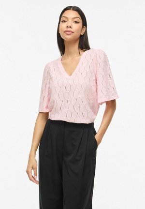 Žena s dlhými tmavými vlasmi, oblečená v svetloružovej textúrovanej blúzke s výstrihom do V a čiernych vysokých nohaviciach, ruky vreckách.