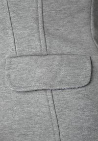 Grått tyg med en slät textur, som har en framficka med veck och vertikala sömmar. Sömmarna är synliga längs kanterna.