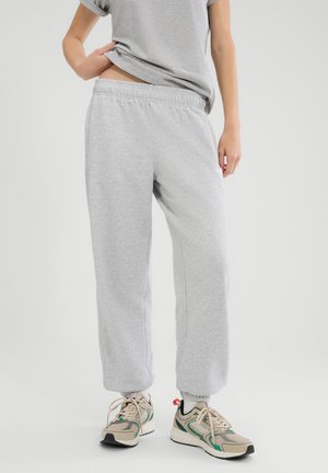 Person iført lysegrå joggingbukser, matchende grå T-shirt og beige sneakers med grønne og røde detaljer på en ensfarvet baggrund.