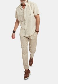 Beige košile s krátkým rukávem a jemným textovým vzorem, spárovaná s světle béžovými kalhotami a hnědými botami. Mezi zajímavé detaily patří kapsa na prsou a ohrnuté manžety.