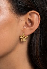 Goldfarbene, blumenförmige Ohrringe mit strukturierten Blütenblättern und einer zentralen Perle, vor einem Nahaufnahme eines Ohres, das glatte Haut und dunkles Haar zeigt.