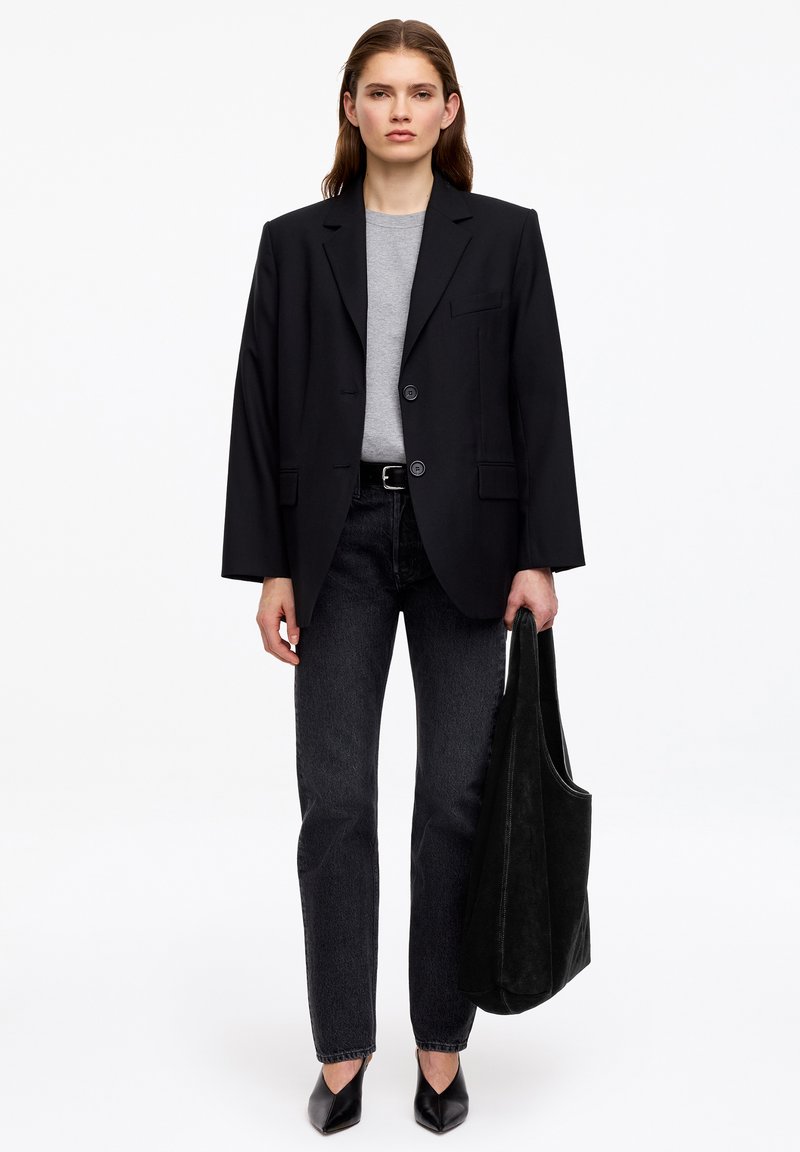 Femme portant un blazer noir oversize, chemise grise, jean noir, chaussures noires pointues, tenant un grand sac cabas noir sur fond blanc uni.