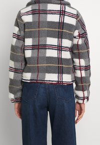 Fleece bunda v šedém károvaném vzoru s bílými, červenými a žlutými proužky; zkrácený střih s límcem; texturovaný povrch a široké rukávy. Viditelné džíny.