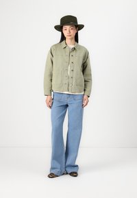Õlivärvi kanvasjakk suurte taskutega, millele on lisatud helesinised flared-jeans. Musta ja pruuni leopardimustri kingad. Lai äärega roheline müts.
