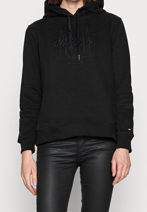 Osoba w czarnej bluzie z kapturem z wytłoczonym logo Tommy Hilfiger i czarnych spodniach z ekologicznej skóry na jednolitym jasnym tle.