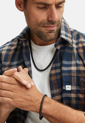Zwarte ketting en armband op een man. Hij draagt een blauw geruit overhemd over een wit T-shirt. De focus ligt op de metalen textuur en het contrast.