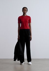 Piros rövid ujjú felső hímzett szöveggel, fekete bő fazonú melegítőnadrággal párosítva. Fehér tornacipő és a bal kézben tartott fekete dzseki.