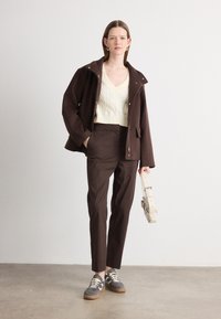 Brown cropped jacket with snaps, cream cable-knit sweater, brown trousers, and gray sneakers with gum soles. Holding a beige shoulder bag.