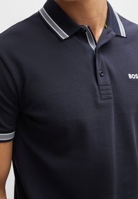 Marinblå piké med texturerad tyg, utrustad med en kontrasterande randig krage, tre knappar och en liten logotyp på bröstet.