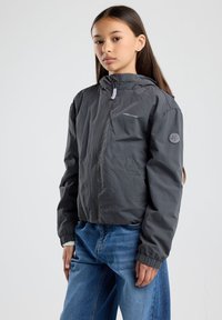 Hameen harmaa kevyt takki, jossa on edessä vetoketju ja huppu, vasemmassa hihassa logo-patch ja elastiset hihansuut. Pukeutunut sinisiin farkkuihin.