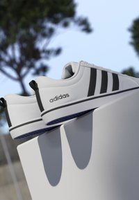 Zapatillas blancas Adidas con rayas negras y logo, colocadas sobre una superficie blanca al aire libre con árboles desenfocados y cielo azul al fondo.