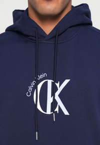 Tmavomodrá mikina s kapucňou z bavlnenej zmesi, s bielym logom Calvin Klein a grafikou "CK". Obsahuje nastaviteľné šnúrky a predné vrecko.