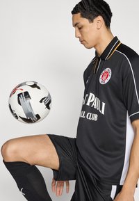 Μαύρη ποδοσφαιρική φανέλα με λευκές λεπτομέρειες και το λογότυπο του συλλόγου, συνδυασμένη με μαύρο σορτς. Μία ποδοσφαιρική μπάλα με σχέδια ισορροπεί στο γόνατο του παίκτη.