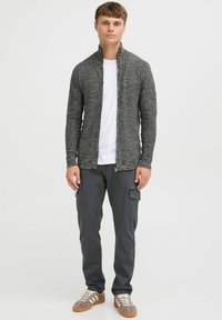 Cardigan de punto gris con cuello alto y cierre de cremallera, usado sobre una camisa blanca; combinado con pantalones cargo oscuros y zapatillas beige.