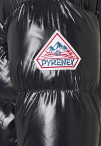 Svart puffjacka med en glänsande, slät yta. Har en framträdande triangelformad Pyrenex-logga i rött, vitt och blått framtill.