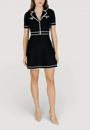 Donna che indossa un vestito nero a maniche corte con bordi bianchi, bottoni e tacchi alti beige, in piedi contro uno sfondo chiaro e uniforme.