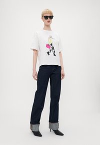 Hvit T-skjorte med en grafisk illustrasjon, matchende mørkeblå jeans med denimmansjetter, og svarte spisse hæler. Svarte solbriller på.