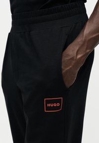 Dunkelhäutige Hand, die in der Seitentasche einer schwarzen Hose mit einem orange umrandeten HUGO-Logo nahe der Tasche ruht.