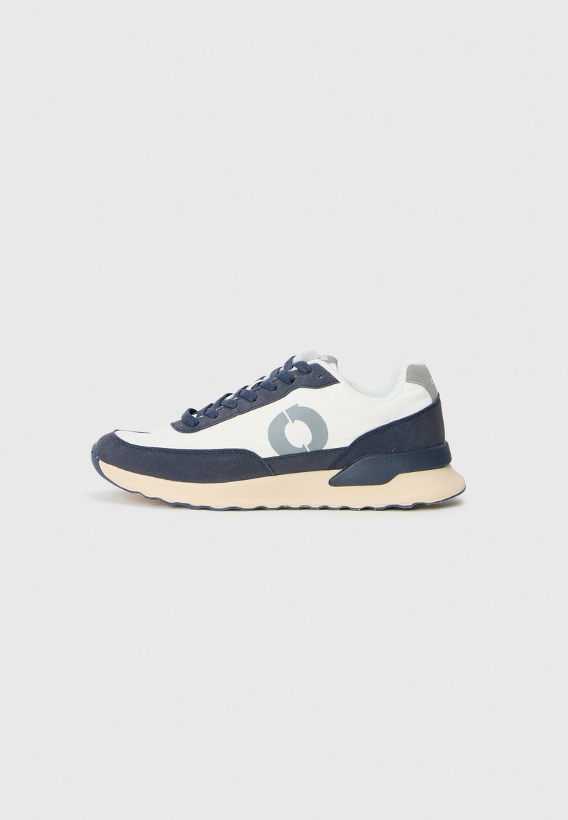 Zapatilla blanca y azul marino con parte superior de ante y malla, puntera redonda, cordones a tono y suela de goma beige. Presenta un logo circular en el lateral.