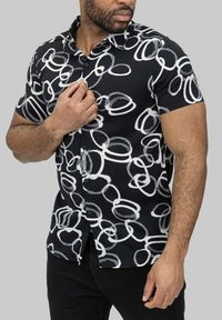 Chemise à manches courtes en tissu noir avec un motif circulaire abstrait blanc et un col classique. Boutons noirs unis visibles.