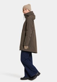 Woman in a brown hooded coat, beige knit hat and gloves, wide-leg blue jeans, and black boots standing in profile on a white background.