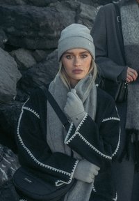 Grijze gebreide beanie, grijze sjaal, grijze handschoenen, zwarte jas met witte borduring. Model staat tegen een donkere rotsachtige achtergrond.