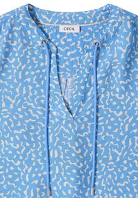 Blouse à motifs bleus et blancs avec col en V, longs cordons bleus avec embouts métalliques, et étiquette de marque "CECIL" à l'intérieur du col.