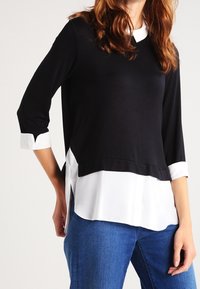 Svart stickad topp med trekvartslånga ärmar, vit skjortsidskänning och kontrasterande vit krage. Mjuk tyg med slät textur.