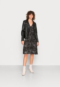 Vestido preto com padrões e mangas curtas, combinado com uma jaqueta de couro preta estilo motociclista. A modelo usa botas de tornozelo em bege claro. Texturas suaves visíveis.