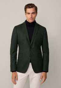 Mörkgrön blazer i texturerat tyg med två knappar, slagkrage och framfickor, bärs över en svart polotröja.