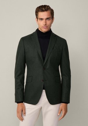 Blazer vert foncé en tissu texturé avec deux boutons, revers crantés et poches avant, porté sur un col roulé noir.