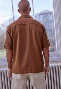 Brązowa teksturowana koszula z krótkim rękawem i kołnierzykiem, zestawiona z jasnymi spodniami, widoczna od tyłu na tle okna.