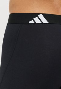 Cinturilla atlética negra con el logo blanco de Adidas; textura ajustada en los pantalones cortos, con detalle de costura visible a lo largo del lado.