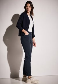 Cárdigan azul marino sobre una camiseta blanca, combinado con pantalones oscuros y zapatillas beige. El conjunto mezcla tejido texturizado con un ajuste casual.