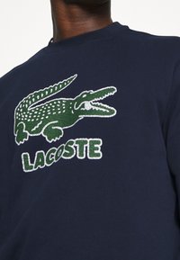 Námořnicky modrá mikina s texturovaným zeleným krokodýlím logem a bílým nápisem "LACOSTE". Kulatý výstřih a měkká textura látky.