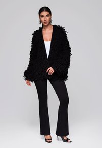 Svart, lurvig cardigan med en lös passform, som har långa, texturerade ärmar och en öppen framsida, bärs över en vit topp och svarta utsvängda byxor.