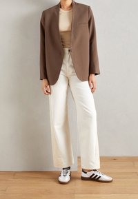 Blazer marrón sobre una blusa beige, combinado con pantalones anchos crema y zapatillas blancas con rayas negras. Diseño sencillo, ajuste moderno.