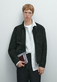 Schwarze Wildlederjacke, weißes kariertes Hemd, dunkle Hose. Hält ein Notizbuch und Bücher. Einfaches Design, mit Reißverschluss, Taschendetails.