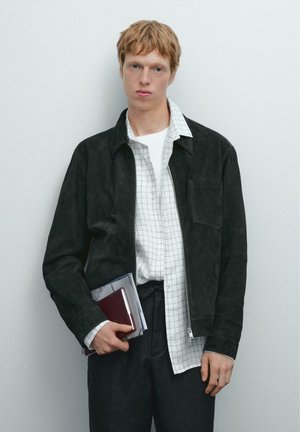 Veste en suede noire, chemise à carreaux blancs, pantalons sombres. Tenant un carnet et des livres. Design simple, fermeture zippée, détails de poche.