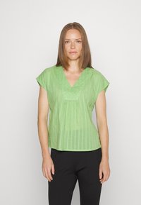 Blusa de manga curta em verde claro, com decote em V, apresentando riscas verticais e um corte ligeiramente descontraído. Usada com calças pretas.