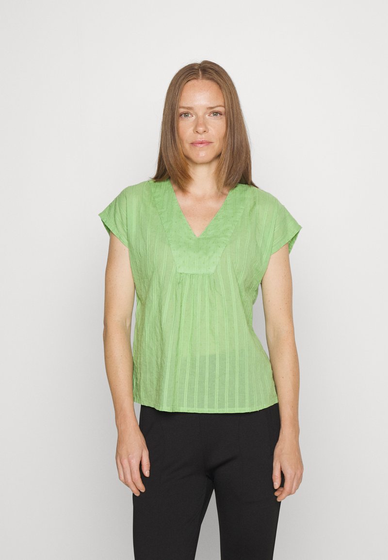 Blusa de manga curta em verde claro, com decote em V, apresentando riscas verticais e um corte ligeiramente descontraído. Usada com calças pretas.