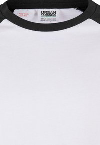 Fehér pamut póló fekete raglán ujjal. Bordás nyakkal rendelkezik, és egy címke található rajta, amelyen ez áll: "Urban Classics, Bangladesben készült."