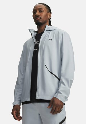Man draagt een lichtgrijze jas met rits en Under Armour-logo, zwart shirt en bijpassende lichtgrijze broek, kijkt zelfverzekerd vooruit.