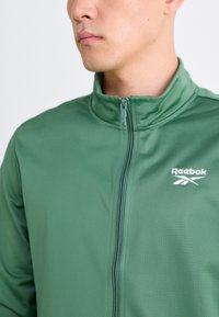 Casaco verde com fecho éclair feito de tecido elástico. Possui um colarinho alto, ajuste slim e um logo branco da Reebok no lado esquerdo do peito.