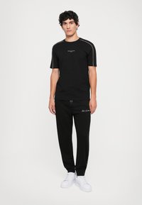 Svart t-skjorte med korte ermer og lys grå detaljer; kombinert med svarte bukser med snøring. Båret med hvite joggesko.