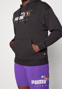 Felpa nera con tasca a marsupio, che presenta il logo multicolore "PUMA LOVE WINS". Abbinata a pantaloni corti viola, anch'essi con il logo PUMA.