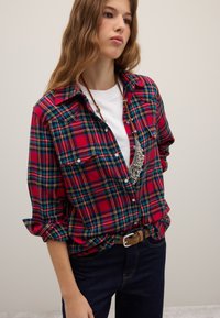 Red and blue plaid flannel shirt with button-down front, rolled sleeves, two chest pockets, worn over a white t-shirt and dark jeans.