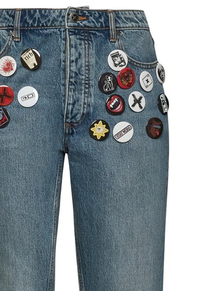 Jeans in denim blu con molteplici spille tonde con vari grafismi e colori attaccate nella parte anteriore, che mostrano una miscela di disegni e textures.