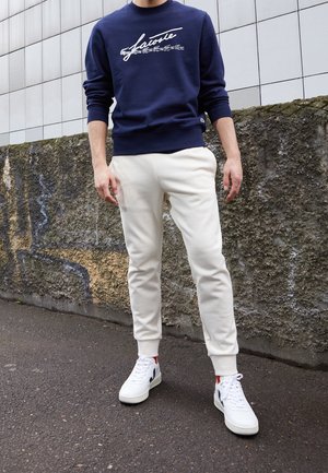 Osoba stojí venku, oblečená v námořnicky modré mikině Lacoste, béžových teplácích a bílých teniskách, na pozadí je strukturovaná kamenná zeď a dlaždicová fasáda.