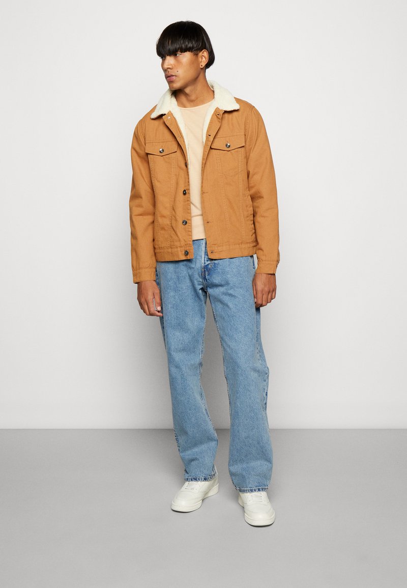 Lichtbruine dennenjas met een schapenvachtkraag, twee borstzakken, gecombineerd met lichtblauwe jeans en witte sneakers. Eenvoudig ontwerp, casual stijl.
